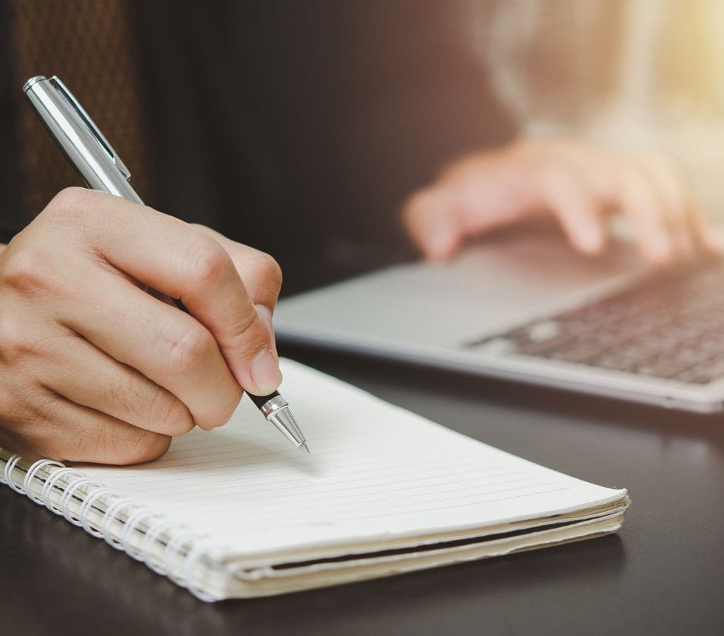 a man writing on a note while typing on his laptop